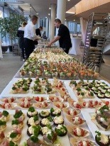 Stół z przekąskami na przyjęciu, widok z góry, różnorodne kanapki i koreczki ułożone w rzędach, obsługa w tle przygotowuje kolejne porcje, jasne oświetlenie sali.