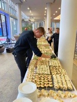 Przygotowywanie różnorodnych przekąsek na długim stole cateringowym, pracownik w czarnych rękawiczkach układa kanapki i roladki, widoczne duże okna i osoby w tle.