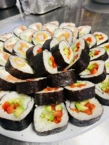 Duży talerz z różnymi rodzajami sushi, w tym maki z łososiem, krewetkami i warzywami, ułożonymi warstwami na białym talerzu, z widocznym fragmentem metalowego blatu w tle.