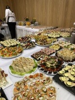 Stół z różnorodnymi przekąskami na przyjęciu, widoczne kanapeczki, sałatki, koreczki i inne finger food, w tle osoba z obsługi w białej koszuli.