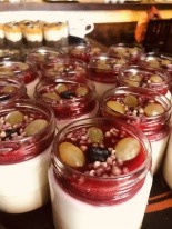Deser w słoiczkach z kremową bazą, warstwą czerwonego dżemu i dekoracją z winogron, orzechów i suszonych owoców, ustawiony w rzędach.