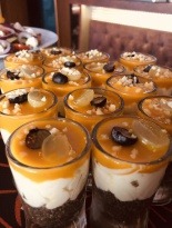 Deser w małych szklankach z warstwą ciemnych okruchów na dnie, białego kremu, pomarańczowej galaretki i kawałkami owoców oraz posypką, ustawiony na stole podczas przyjęcia.