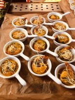 Bufet z małymi porcjami zupy krem z krewetką i posypką, podane w białych ceramicznych naczyniach w kształcie łyżek, na tle brązowego papieru.