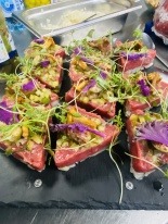 Tatar wołowy ułożony na kwadratowych grzankach, udekorowany kiełkami i fioletowymi płatkami na czarnym, kamiennym talerzu.