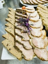 Plater z różnorodnymi wędlinami: pasztet krojony w prostokąty, pieczeń wieprzowa w plastrach, udekorowane gałązką ziół i pomidorkiem cherry na białym talerzu.