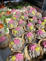 Taca z przekąskami: kanapeczki z pomidorkami cherry i zielenią oraz tartaletki z szarą pastą, różową dekoracją i zielonymi kuleczkami, ułożone w rzędach.