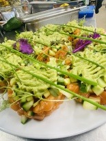 Porcje tatara z łososia z awokado, ogórkiem i szczypiorkiem na białych talerzach, przygotowane w kuchni cateringowej.