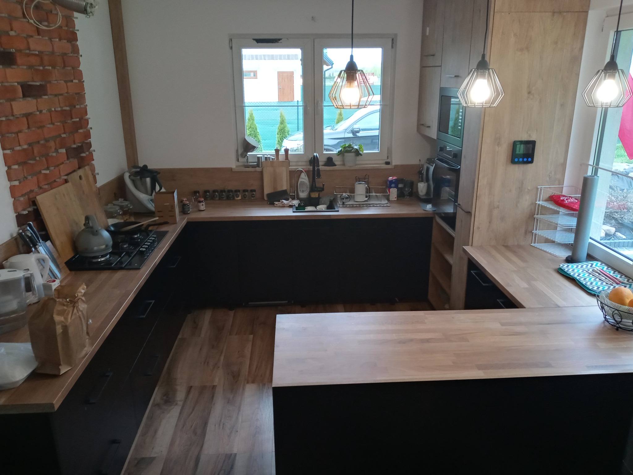 Jasna kuchnia z drewnianymi blatami, czarnymi szafkami i ścianą z czerwonej cegły, oświetlona trzema wiszącymi lampami z żarówkami Edison, widok na zewnątrz przez okno.