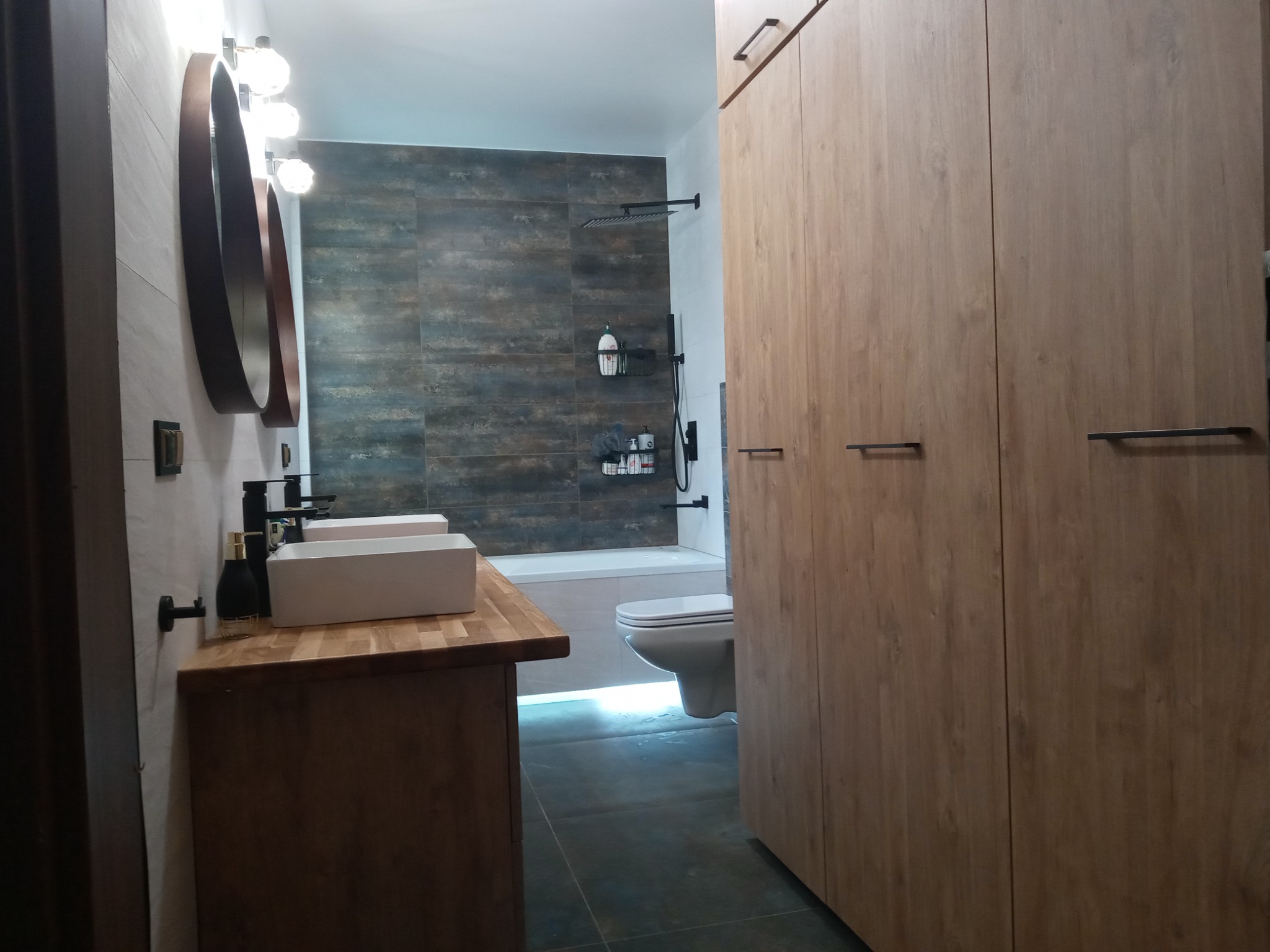 Nowoczesna łazienka z drewnianą szafką pod umywalką z dwoma białymi, prostokątnymi umywalkami, okrągłymi lustrami i dużą szafą z drewnianymi frontami.