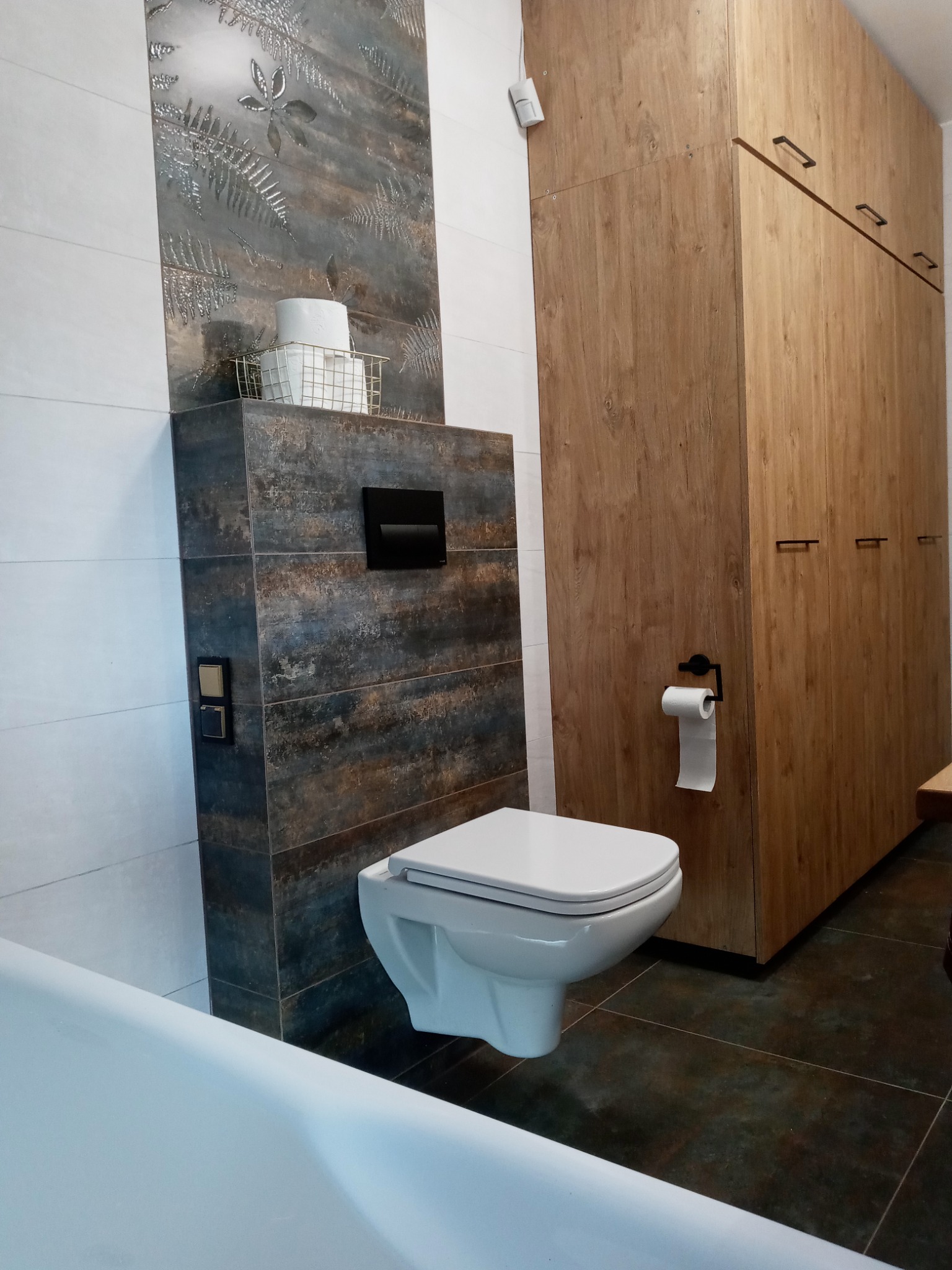 Nowoczesna łazienka z wiszącą toaletą i zabudową meblową z drewnopodobnych płyt, dekoracyjna płytka z motywem roślinnym nad stelażem, widoczna część wanny.
