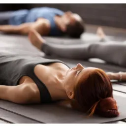 Grupa osób leżących na matach w pozycji relaksacyjnej podczas zajęć. Kobieta z rudymi włosami spiętymi w kok na pierwszym planie, widoczny tatuaż na ramieniu osoby obok.