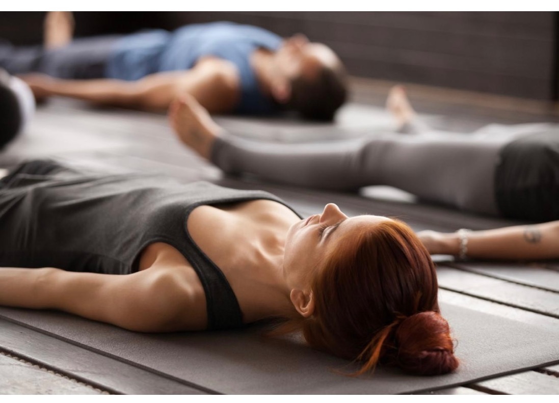 Grupa osób leżących na matach w pozycji relaksacyjnej podczas zajęć. Kobieta z rudymi włosami spiętymi w kok na pierwszym planie, widoczny tatuaż na ramieniu osoby obok.