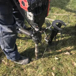 Praca w terenie: Wiertnica udarowa zagłębia rurę w ziemi, operator w ubraniu roboczym stoi obok na trawie.