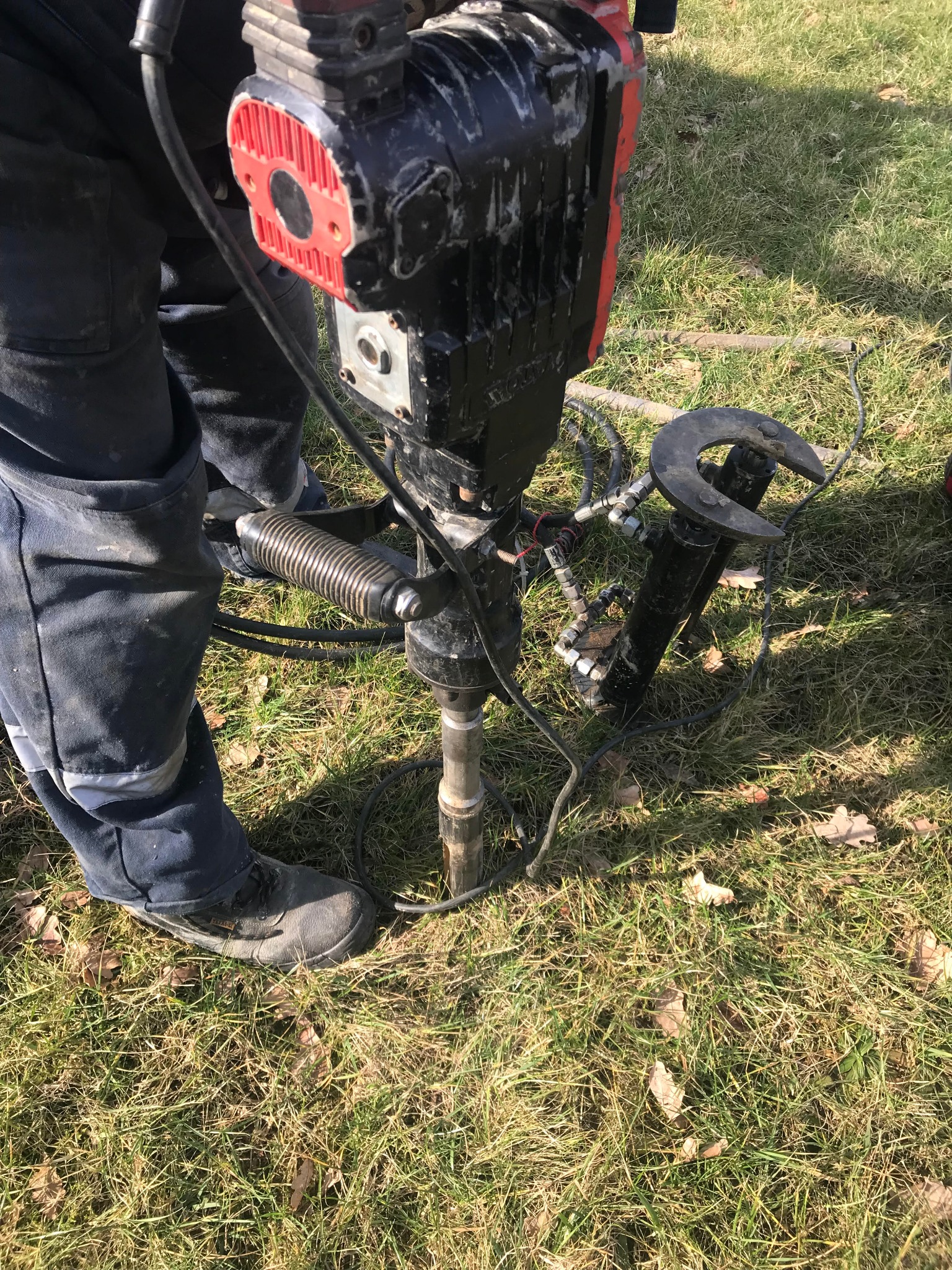 Praca w terenie: Wiertnica udarowa zagłębia rurę w ziemi, operator w ubraniu roboczym stoi obok na trawie.