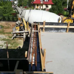 Betoniarka i zbrojenie w szalunku fundamentu płyty betonowej na tle domu z czerwoną dachówką.