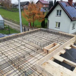 Zbrojenie stropu domu jednorodzinnego z drewnianym szalunkiem, widok z góry na siatkę z prętów stalowych i elementy deskowania, w tle dom z panelami słonecznymi.