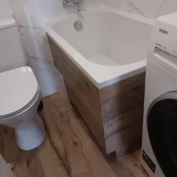 Wykończona łazienka w Żelechowie: biała wanna obudowana panelami imitującymi drewno, biała toaleta i pralka na drewnianej podłodze, ściany wyłożone marmurem.