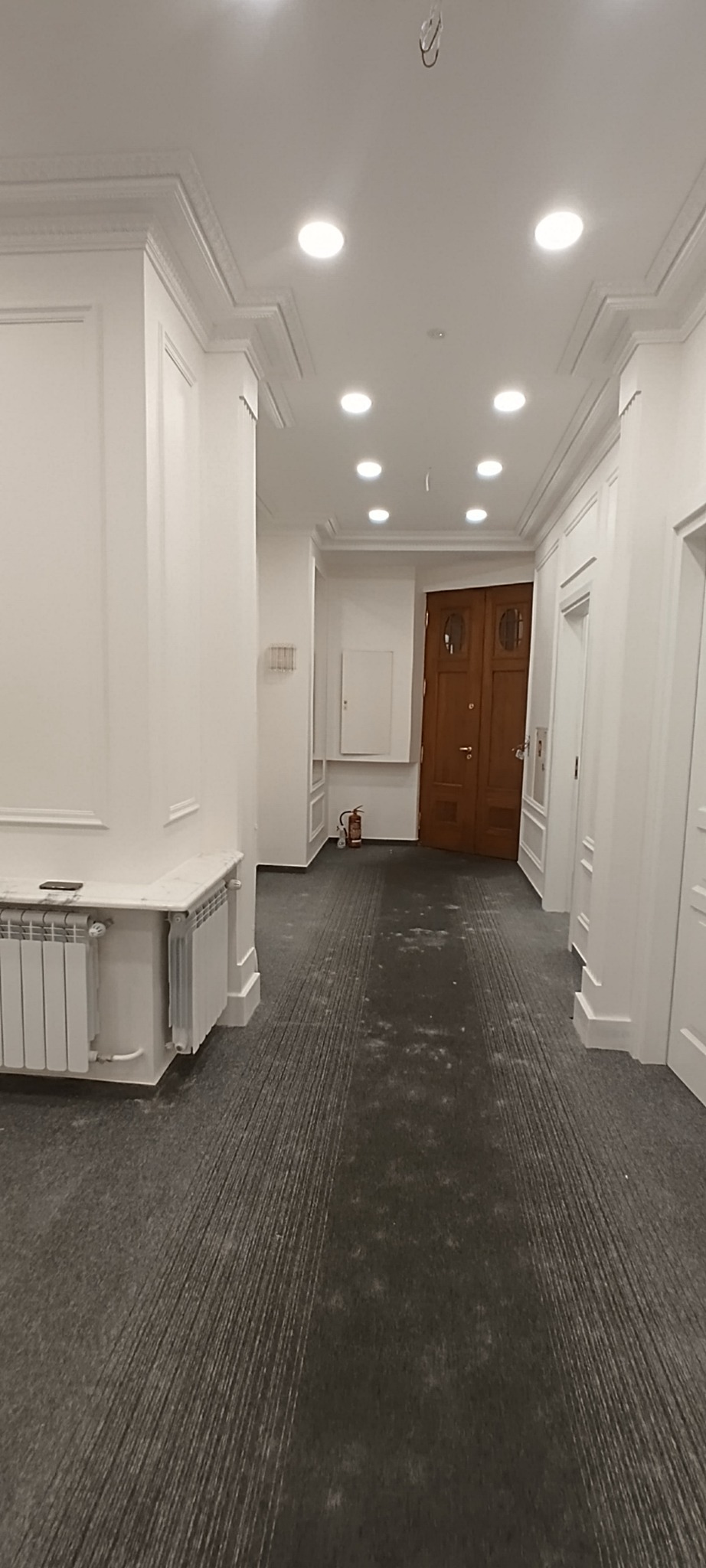 Korytarz biurowy po remoncie z nową szarą wykładziną dywanową, białymi ścianami z dekoracyjnymi listwami i sufitem z oświetleniem punktowym, widoczny grzejnik i brązowe drzwi.