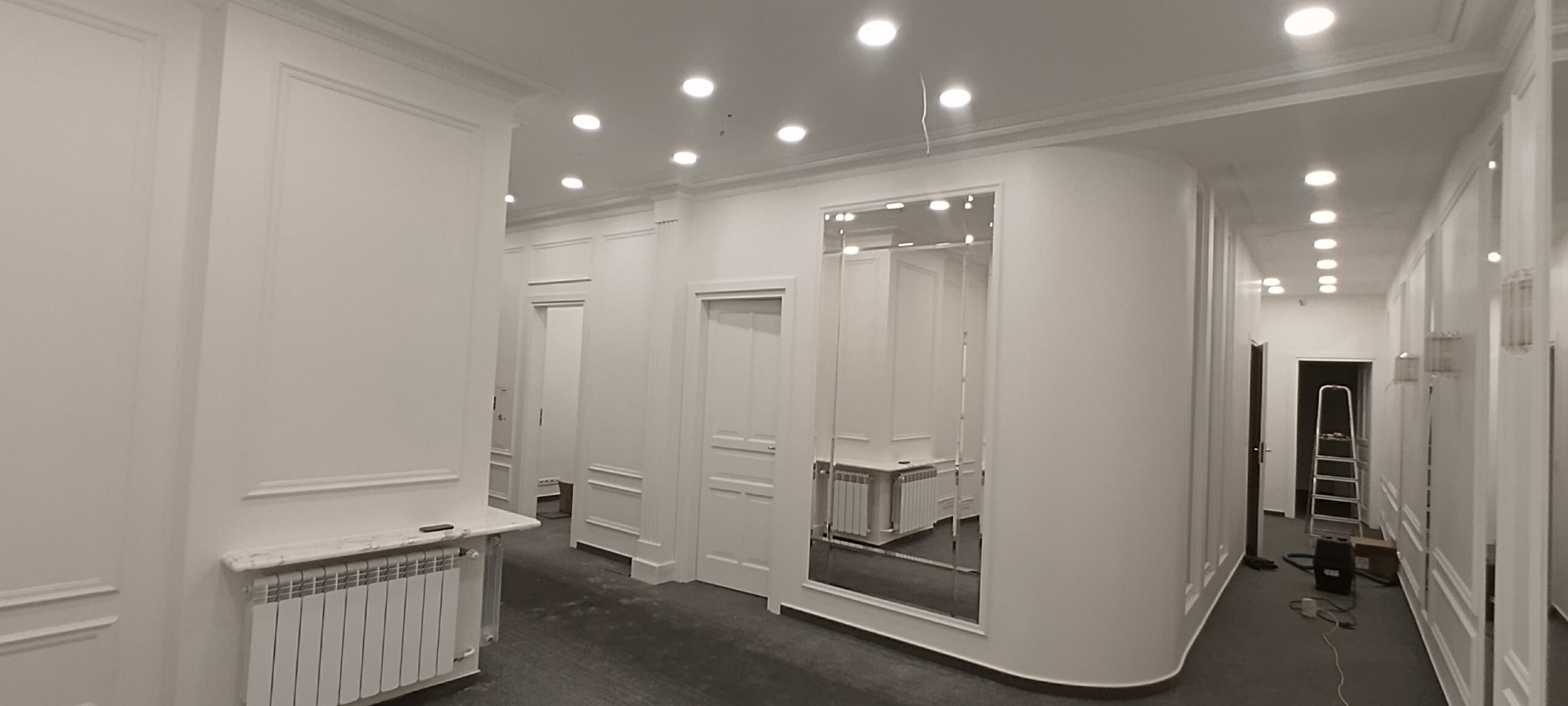 Jasne biuro z białymi ścianami, dekoracyjnymi listwami, oświetleniem punktowym i fragmentem drabiny w głębi korytarza.