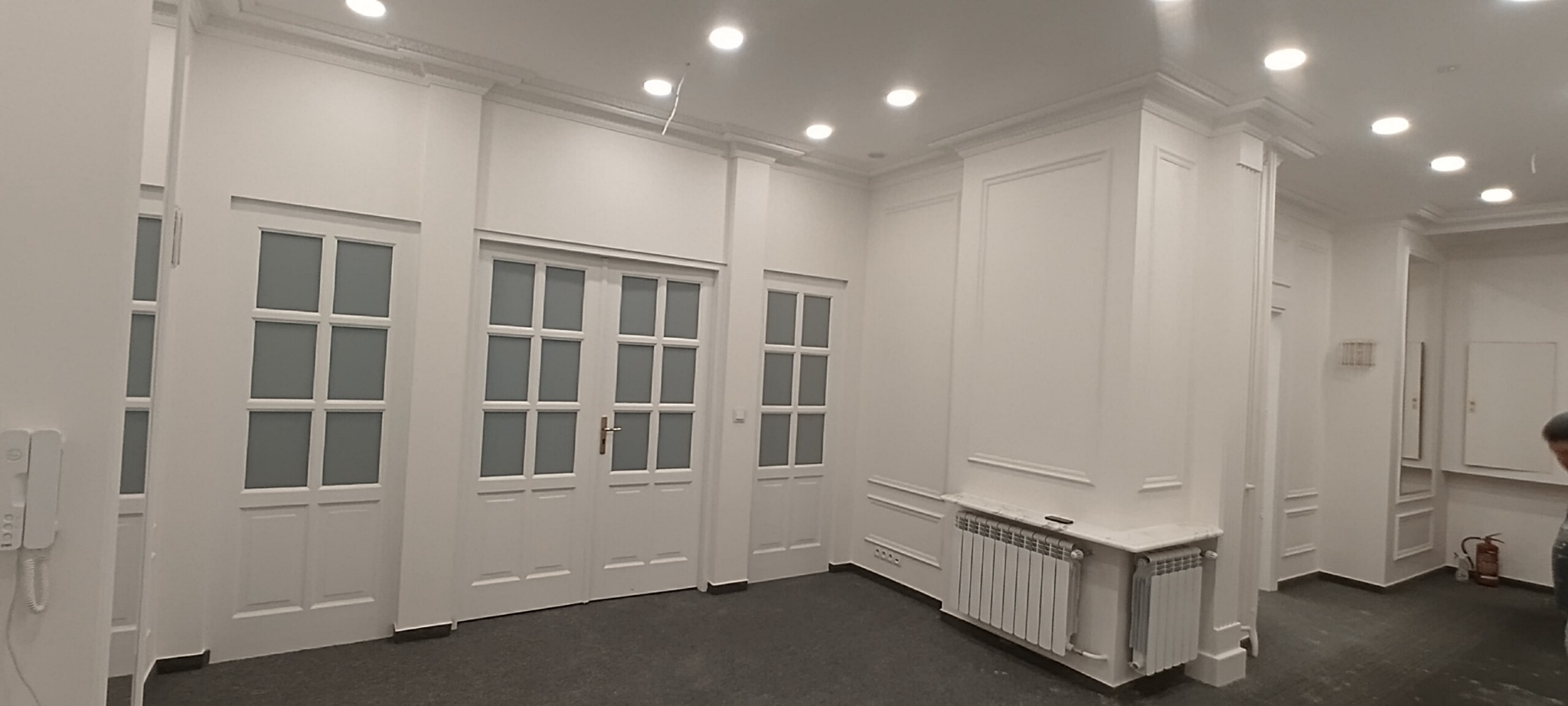 Białe, eleganckie wnętrze biura z trzema parami przeszklonych drzwi, dekoracyjnymi listwami ściennymi i grzejnikami pod marmurowymi parapetami.