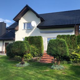 Complex-Dach Dawid Dębski - Dom jednorodzinny z ciemną dachówką, jasną elewacją, widoczną anteną satelitarną i ozdobnymi krzewami w ogrodzie.