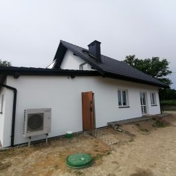 Complex-Dach Dawid Dębski - Nowoczesny, biały dom jednorodzinny z czarnym dachem z blachy trapezowej, widoczny z boku, z otwartymi drewnianymi drzwiami wejściowymi i klimatyzatorem Samsung na ścianie, teren wokół domu...