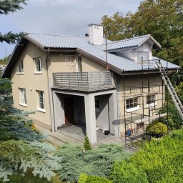 Complex-Dach Dawid Dębski - Dwupiętrowy dom z szarą dachówką, częściowo osłonięty rusztowaniem i folią ochronną podczas prac elewacyjnych, widok z góry na bujną zieleń.
