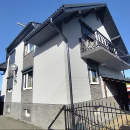 Elewacja dwukondygnacyjnego domu z szarą fasadą, balkonem z ozdobną balustradą, ciemnoszarą stolarką okienną i dachem, widoczny fragment rynny i satelita na ścianie.