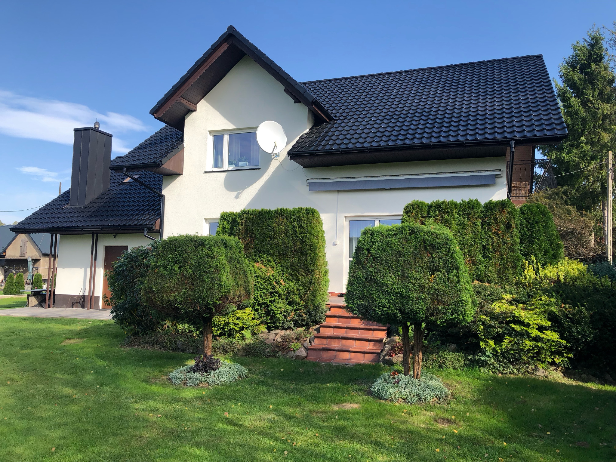 Dom jednorodzinny z ciemną dachówką, jasną elewacją, widoczną anteną satelitarną i ozdobnymi krzewami w ogrodzie.