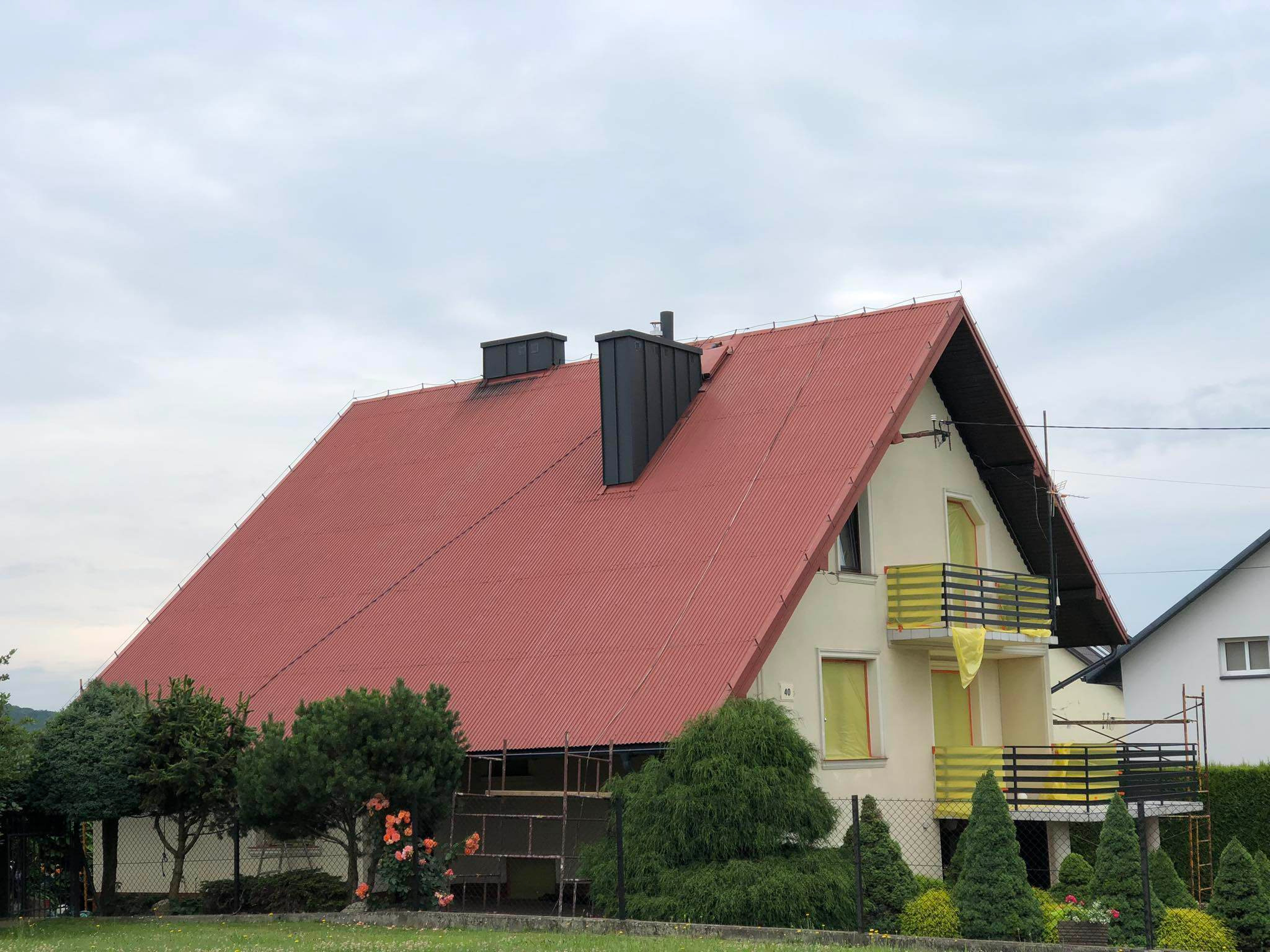 Dom jednorodzinny z czerwonym dachem z blachy trapezowej i dwoma czarnymi kominami, okna i balkony zabezpieczone folią malarską, widoczne rusztowania.