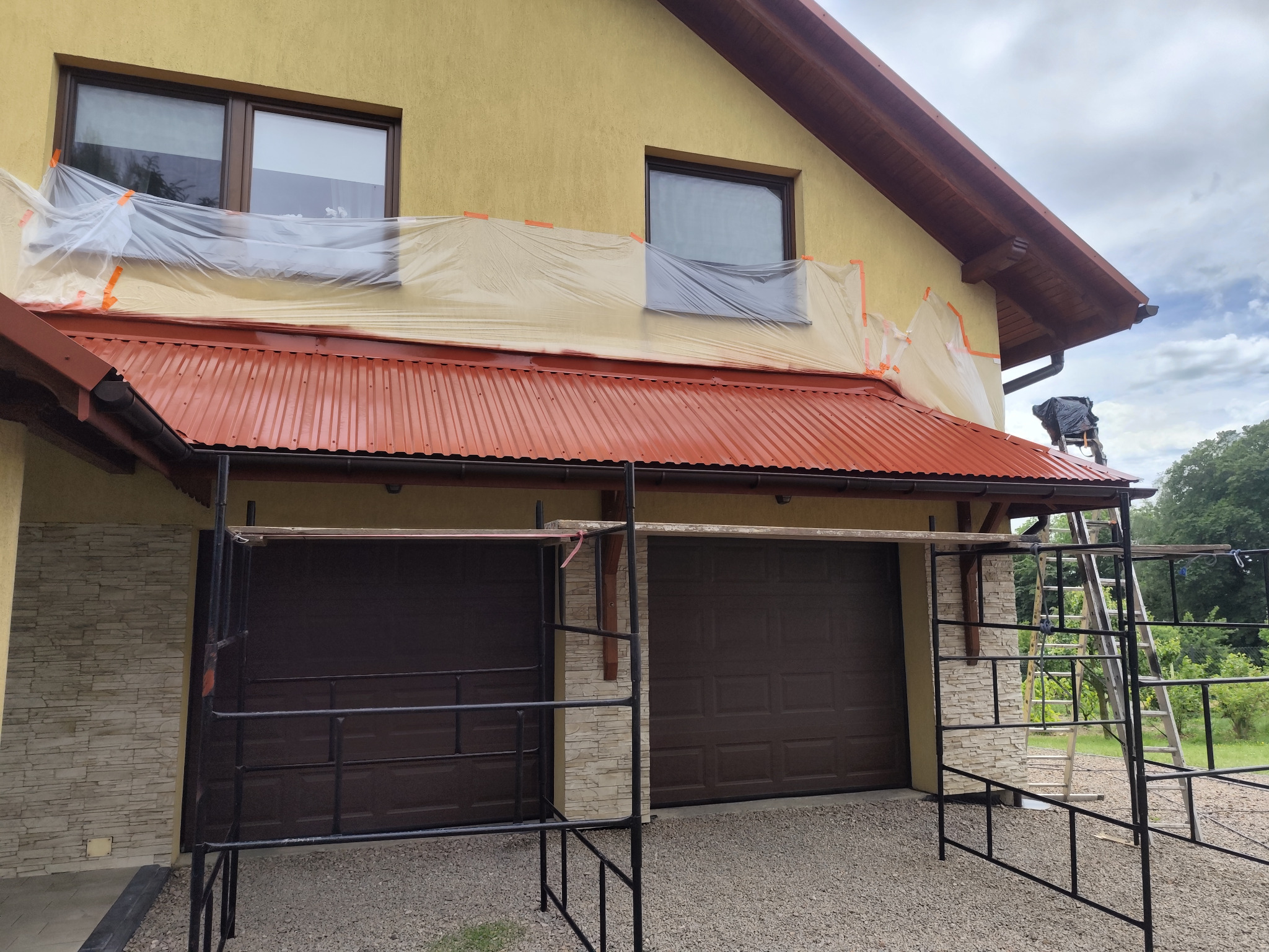 Przygotowania do malowania elewacji domu jednorodzinnego: okna zabezpieczone folią, rusztowanie rozstawione, dach pokryty nową, czerwoną blachą trapezową.