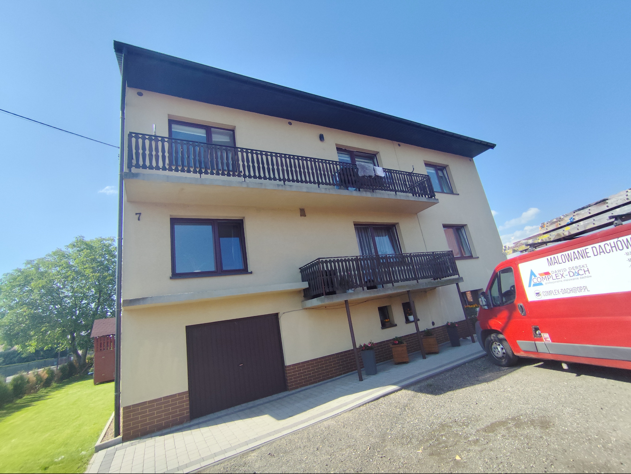 Dwupiętrowy dom z balkonami i ciemną stolarką okienną, z boku zaparkowany czerwony bus z napisem 'Malowanie Dachów' i logo firmy, drabina na dachu busa, błękitne niebo w tle.