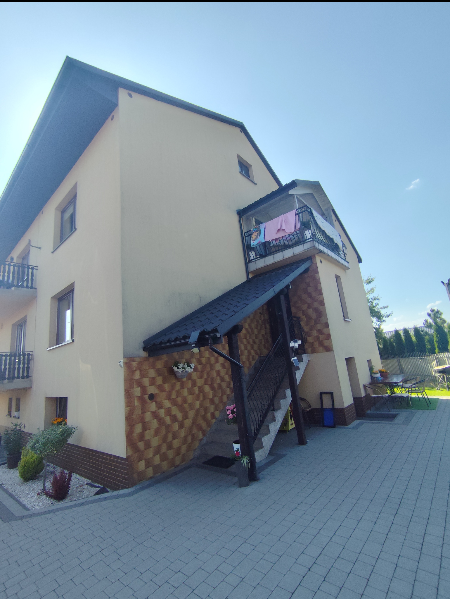 Dwupiętrowy dom z beżową elewacją, ciemnym dachem i balkonem z suszącym się praniem, widziany z dołu pod kątem.