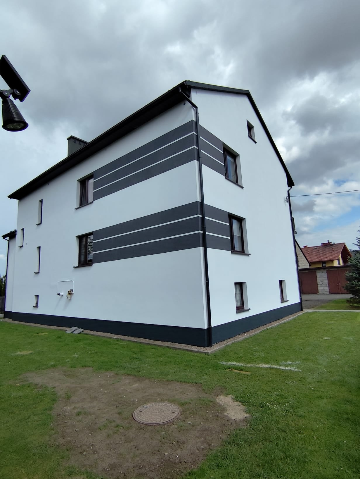 Odnowiona elewacja budynku z białymi ścianami i szarymi pasami, widok z boku na tle pochmurnego nieba.