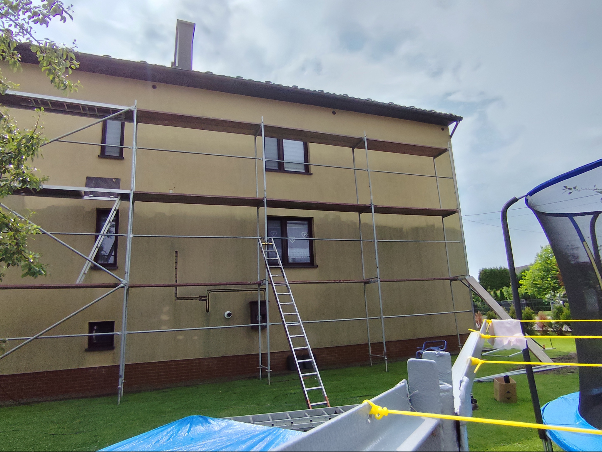 Dom jednorodzinny w trakcie prac elewacyjnych; widoczne rusztowanie, drabina oparta o ścianę, fragment trampoliny w kadrze.