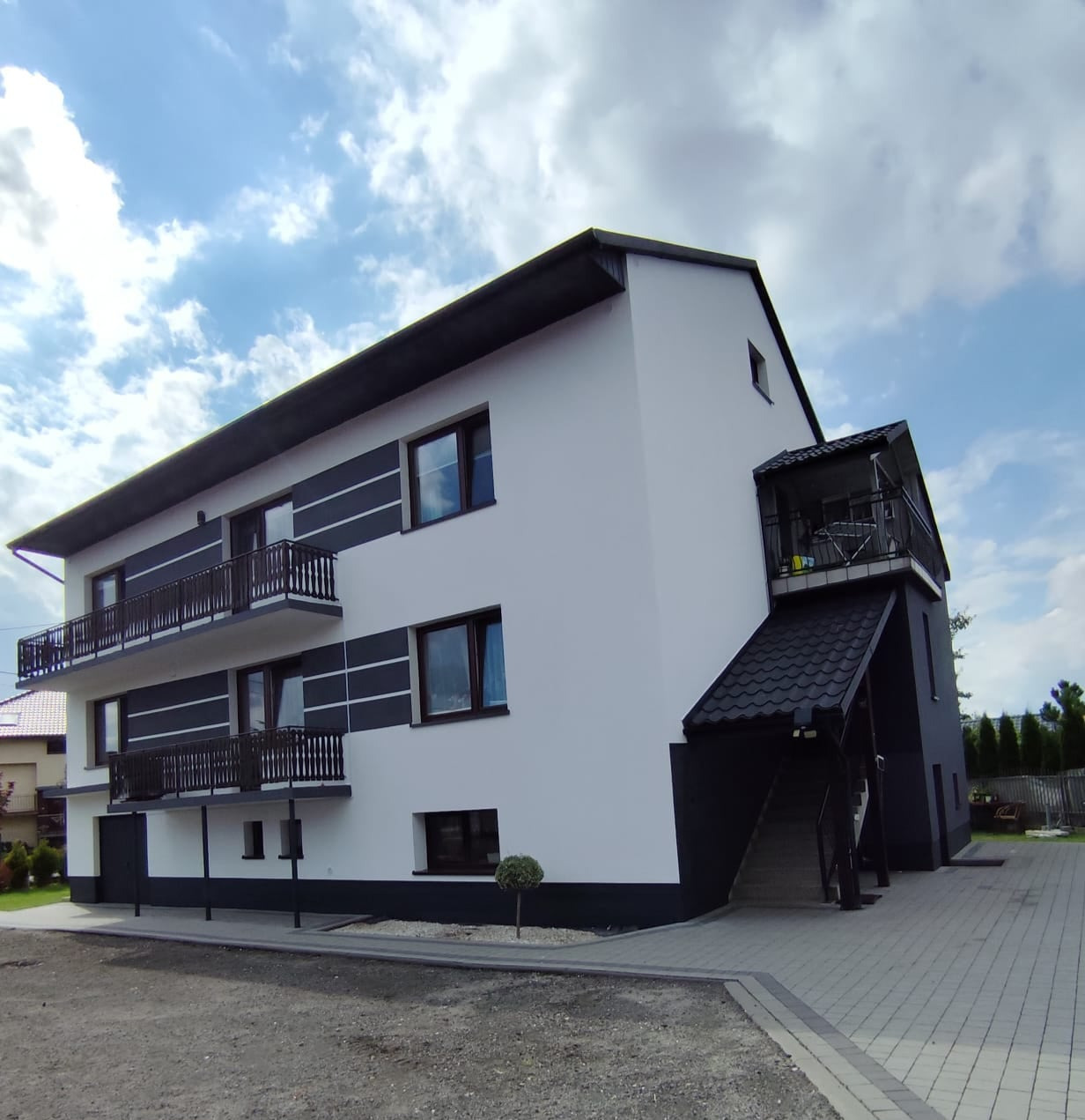 Dwupiętrowy budynek z balkonami i zewnętrznymi schodami, pomalowany na biało z czarnymi akcentami, widok z poziomu gruntu, błękitne niebo w tle.