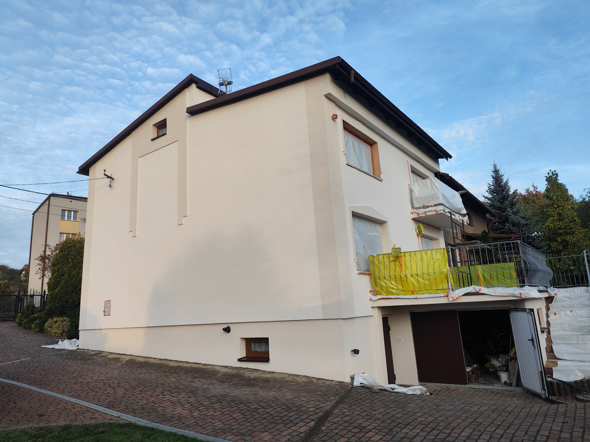 Świeżo pomalowana elewacja domu jednorodzinnego w odcieniach beżu, z zabezpieczonymi oknami i balkonem folią malarską, widoczna kostka brukowa na podjeździe oraz fragment garażu.