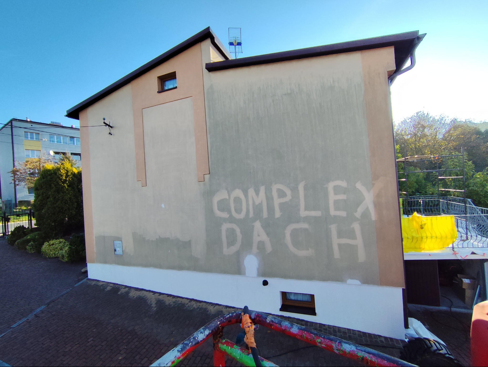Ściana budynku z napisem 'COMPLEX DACH' przed malowaniem, widok z rusztowania, w tle zieleń i inne budynki.