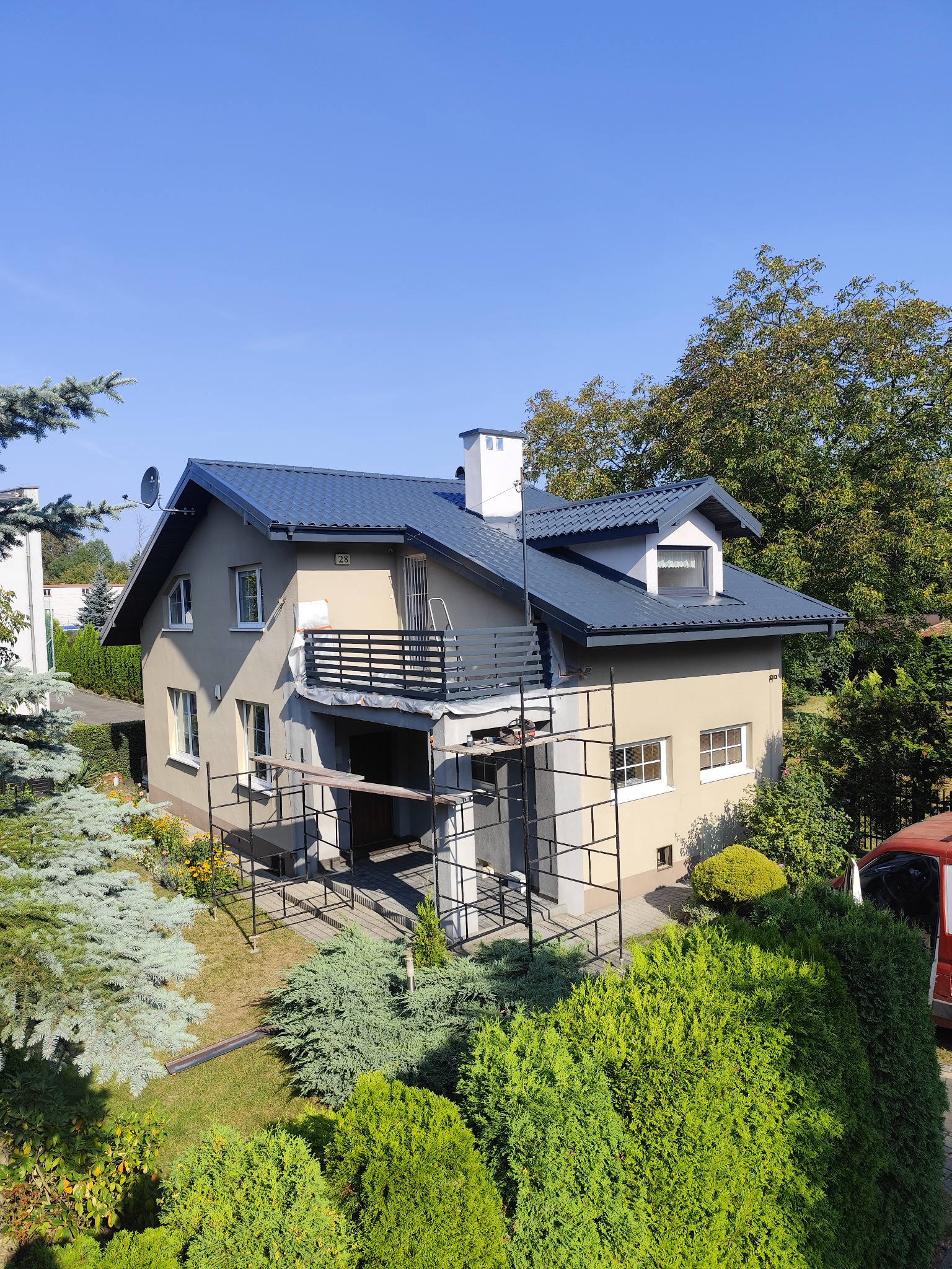 Dom jednorodzinny w trakcie prac elewacyjnych i dekarskich, widoczne rusztowania, odnowiony dach w kolorze grafitowym, jasna elewacja i fragment ogrodu z iglakami.