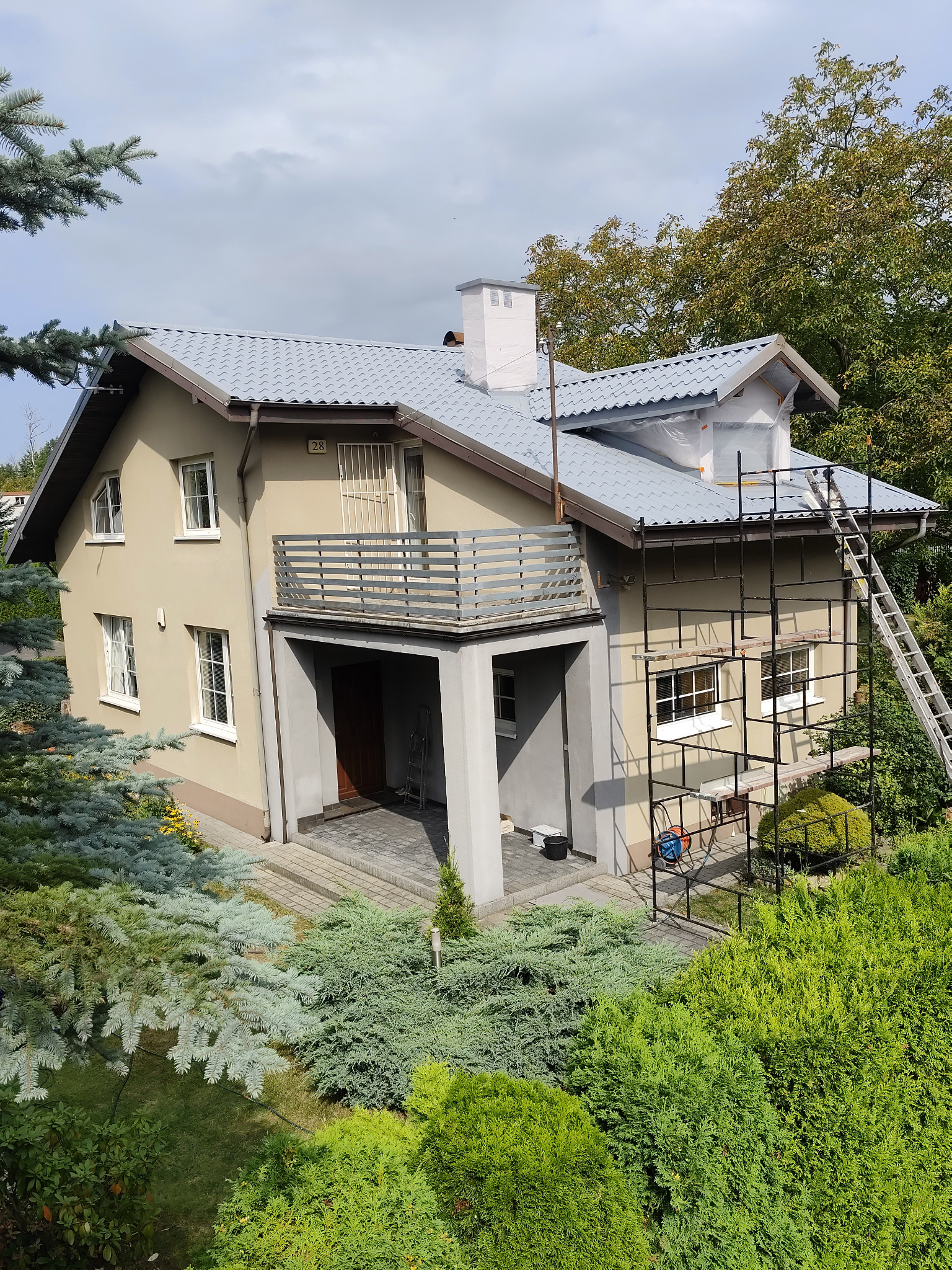 Dwupiętrowy dom z szarą dachówką, częściowo osłonięty rusztowaniem i folią ochronną podczas prac elewacyjnych, widok z góry na bujną zieleń.