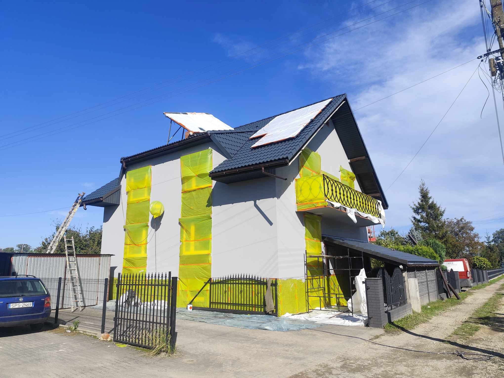 Dom jednorodzinny zabezpieczony żółtą folią malarską przed malowaniem elewacji, widoczna drabina oparta o dach, błękitne niebo w tle.