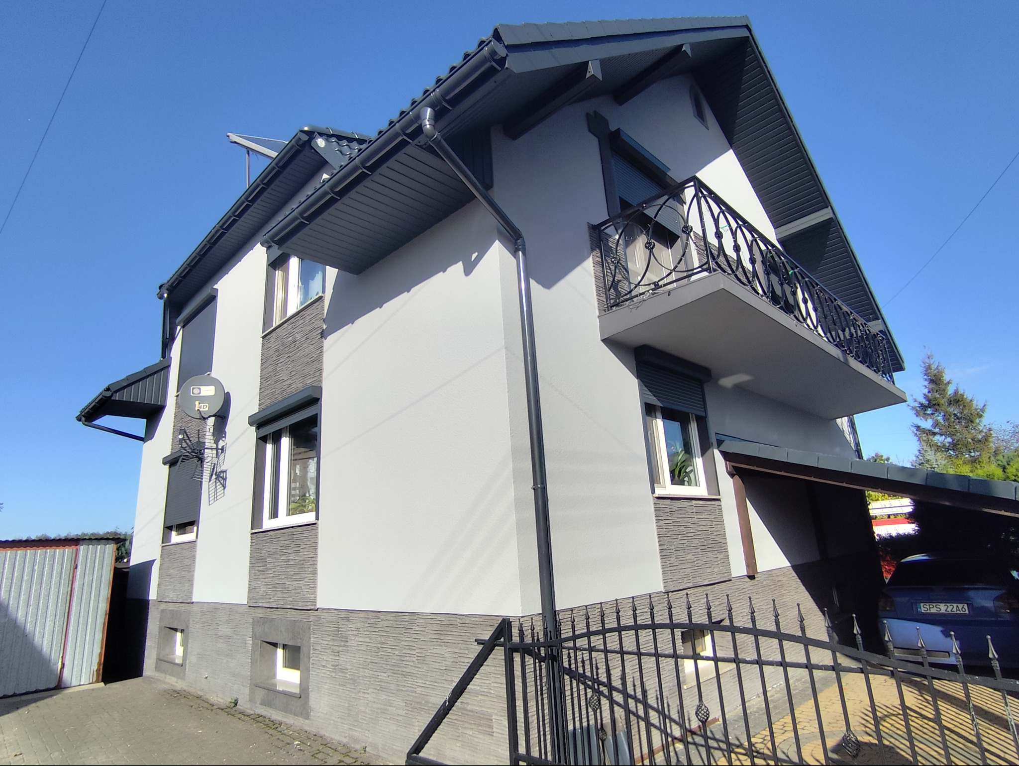 Elewacja dwukondygnacyjnego domu z szarą fasadą, balkonem z ozdobną balustradą, ciemnoszarą stolarką okienną i dachem, widoczny fragment rynny i satelita na ścianie.