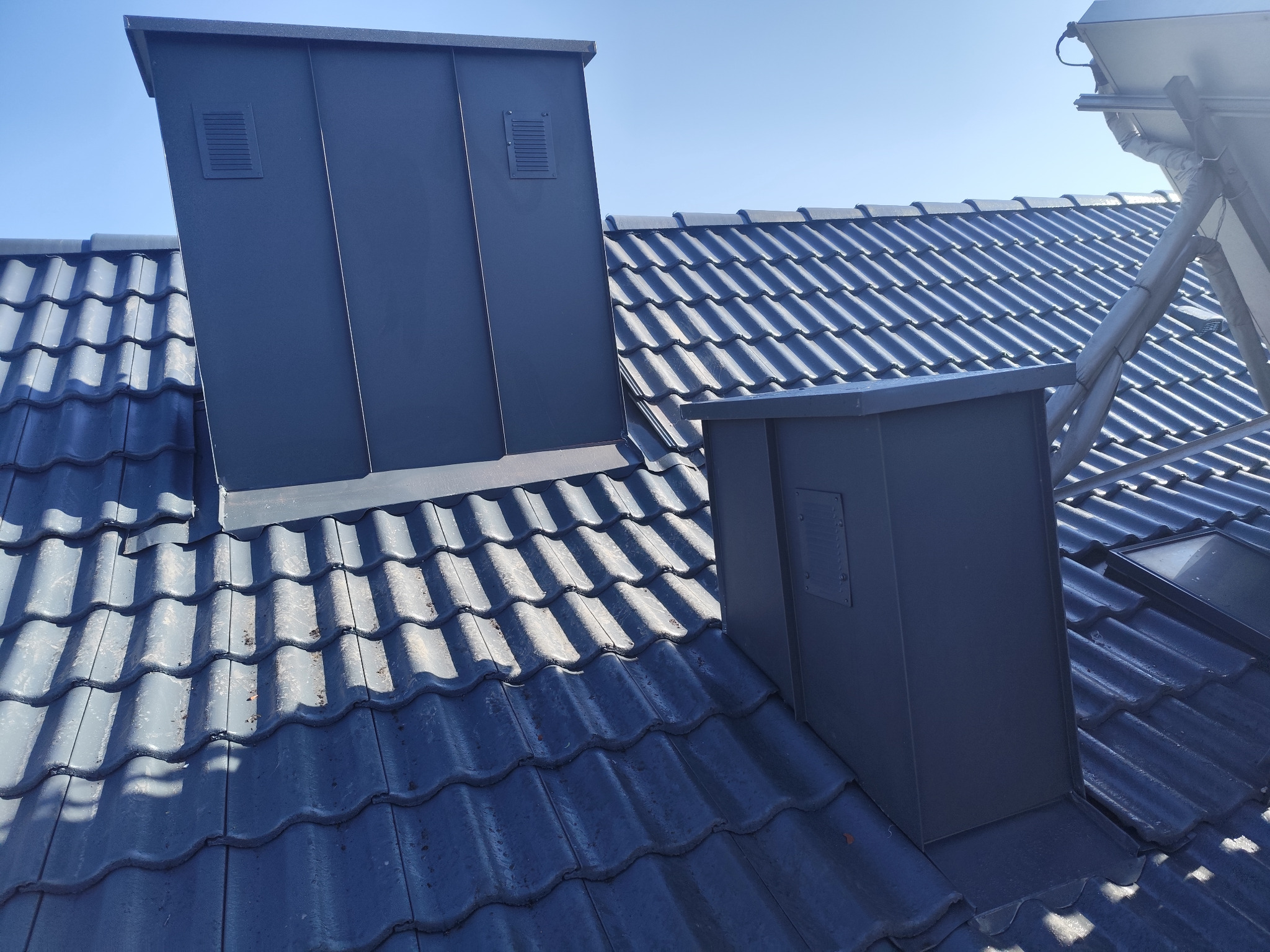 Szare, metalowe obróbki blacharskie kominów na dachu z ciemnej dachówki ceramicznej, widoczne kratki wentylacyjne.