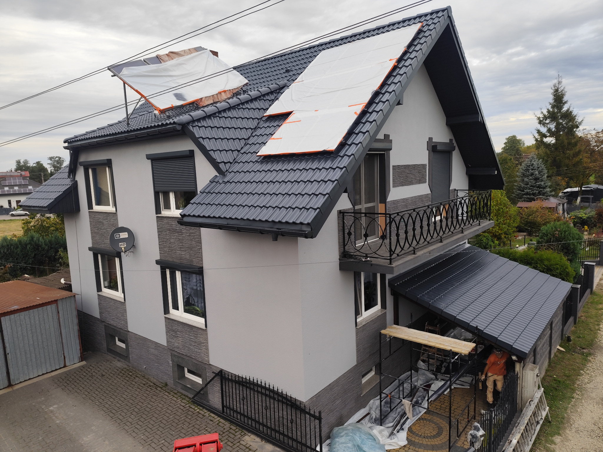 Dwupiętrowy dom z szarą elewacją i ciemnoszarą dachówką, fragment dachu zabezpieczony plandekami, widoczne rusztowanie i pracownik w dolnej części zdjęcia.