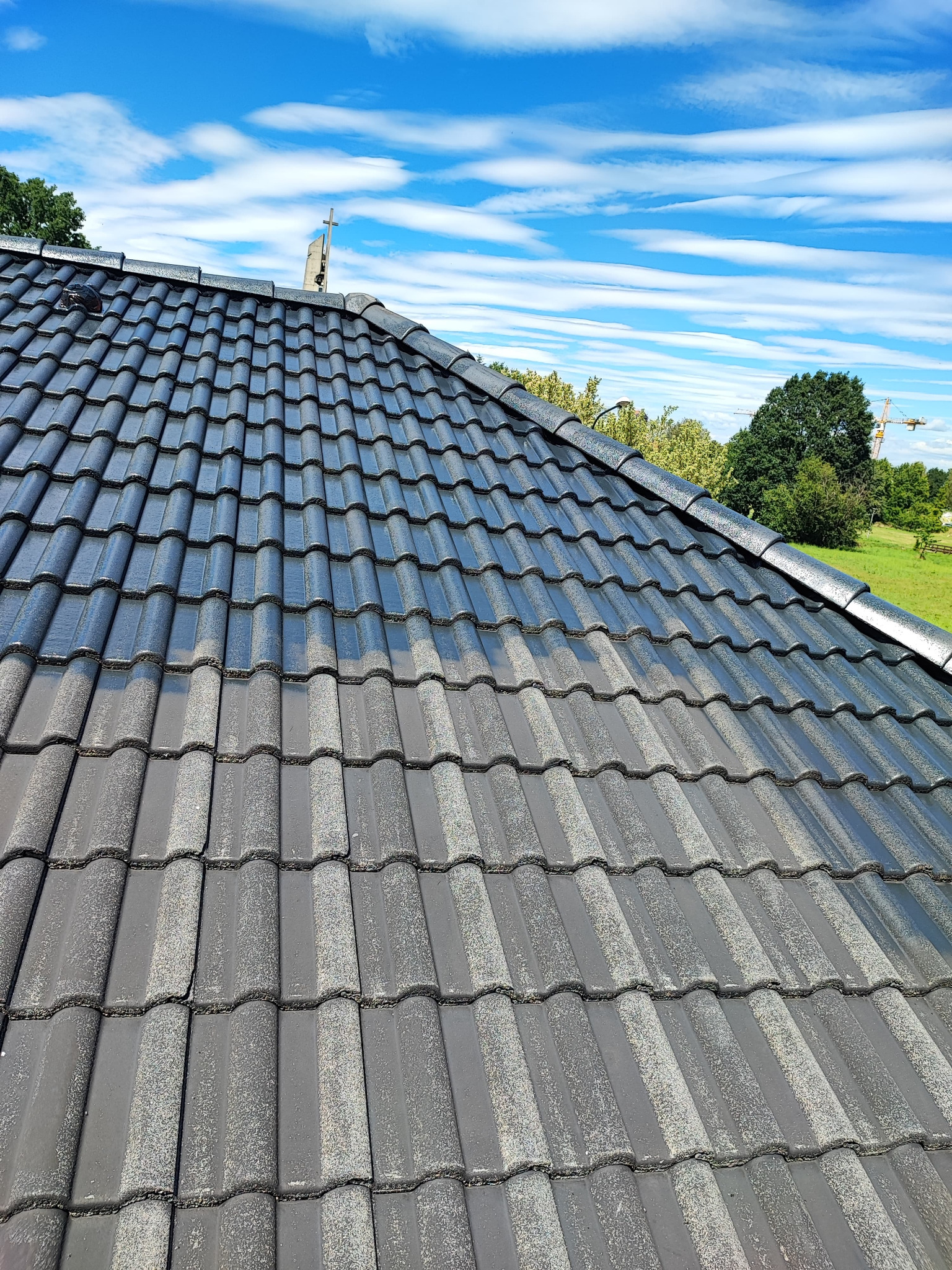 Częściowo umyty dach z szarej dachówki ceramicznej, ukazujący różnicę między czystą a brudną powierzchnią, na tle błękitnego nieba z chmurami.