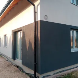 Dwukolorowa elewacja domu jednorodzinnego z widocznym systemem odprowadzania wody deszczowej i fragmentem odsłoniętego fundamentu.