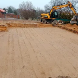 Żółta koparka JCB przygotowuje teren pod przyszły budynek, widoczne wykopane fundamenty i równy piasek, domy w tle.
