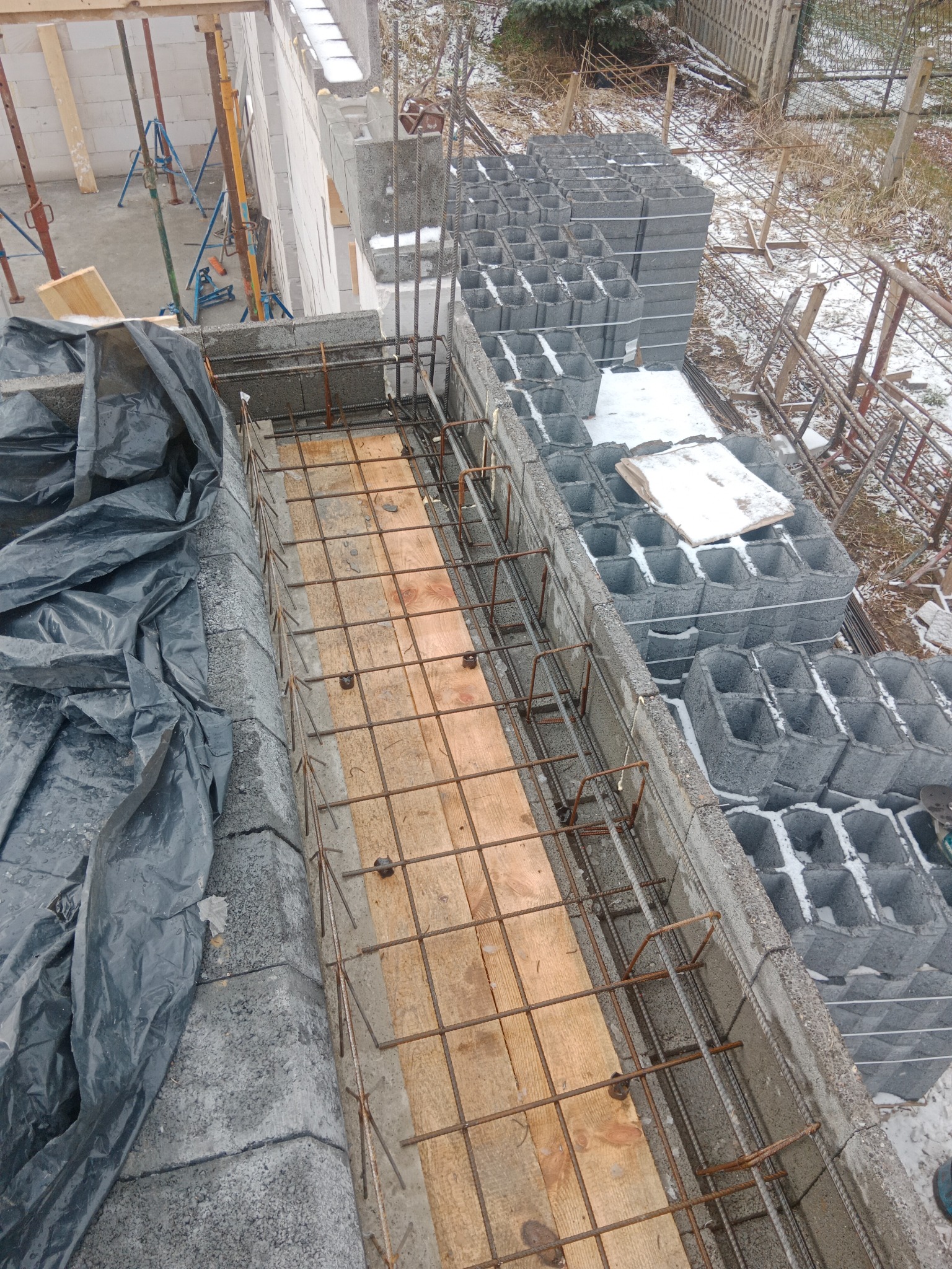 Wykop pod fundament zbrojony stalą, z widocznymi bloczkami betonowymi i deskowaniem, częściowo przykryty czarną folią w zimowej scenerii z resztkami śniegu.