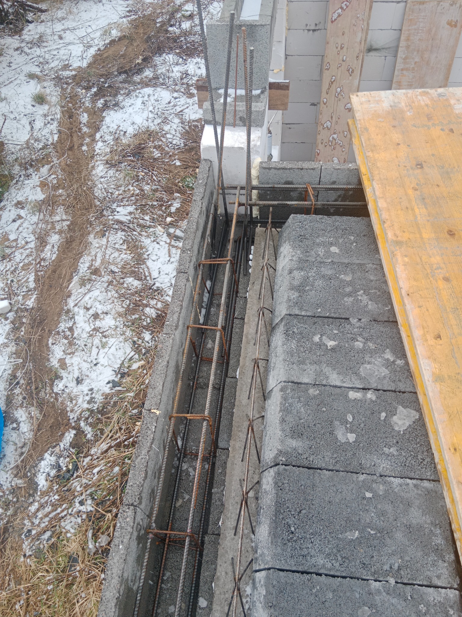 Wznoszenie ściany z bloczków betonowych z widocznym zbrojeniem pionowym i poziomym, częściowo zasypane śniegiem.