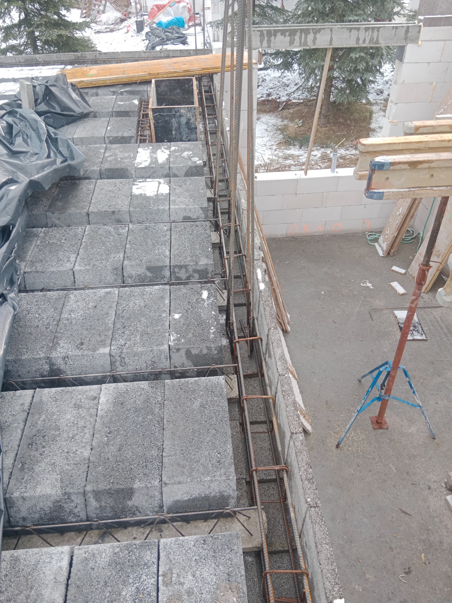 Wznoszenie ścian z szarych bloczków betonowych, zbrojonych stalowymi prętami, widok z góry na budowę w zimowej scenerii.